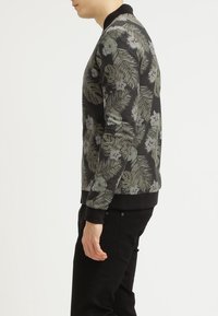 Uomo che indossa pantaloni neri e una camicia a maniche lunghe con un motivo floreale nero, in piedi in profilo contro uno sfondo bianco semplice.