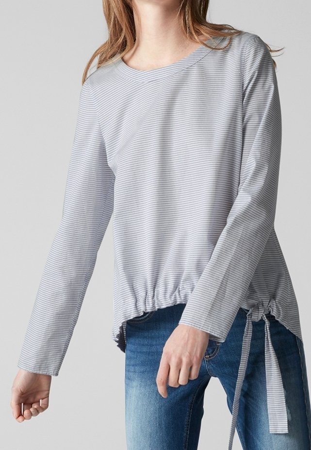 Femme portant un haut à manches longues rayé gris clair et blanc avec un cordon de serrage, associé à un jean en denim bleu.
