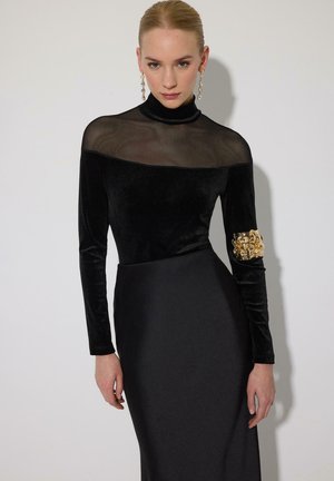 Ženska v črni obleki z visokim prosojnim ovratnikom in zlatim cvetličnim okrasjem na desnem rokavu, stoji pred enakomerno svetlo podlago.