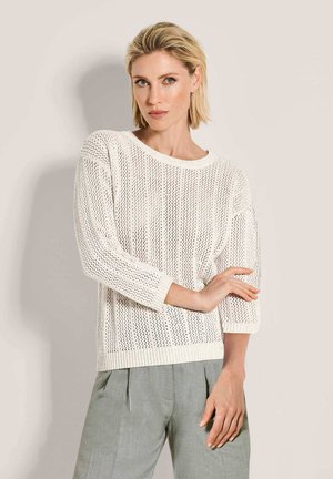 Blonde vrouw die een witte gebreide trui met driekwartmouwen en lichtgrijze plissébroek draagt, poseert met één arm gekruist.