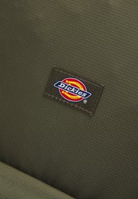Tela de color verde oliva con una superficie texturizada que presenta un parche rectangular del logo de Dickies con acentos en rojo, amarillo y azul.