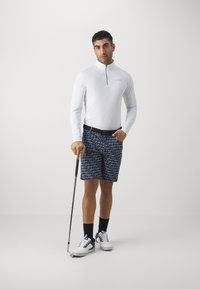 Hvit skjorte med lange ermer og glidelås, mønstrete blå shorts med "Calvin Klein"-tekst, svart belte, golfkølle, hvite sko med svarte detaljer.