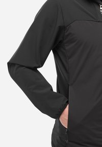 Personne portant une veste noire à manches longues avec une main partiellement dans une poche latérale zippée, le pouce visible reposant à l'extérieur de la poche.