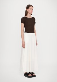 Hnědý pletený top s krátkým rukávem v kombinaci s bílou, volnou maxi sukňou. Modelka má na sobě černé platformové sandály. Minimalistický design a textury.