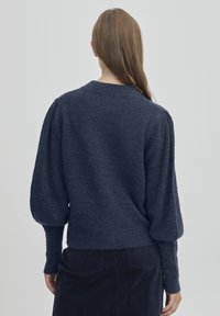 Marinblå stickad tröja med puffärmar och rund halsringning. Mjuk textur, ribbad mudd och nederkant, bärs med mörka kordfloddsbyxor.
