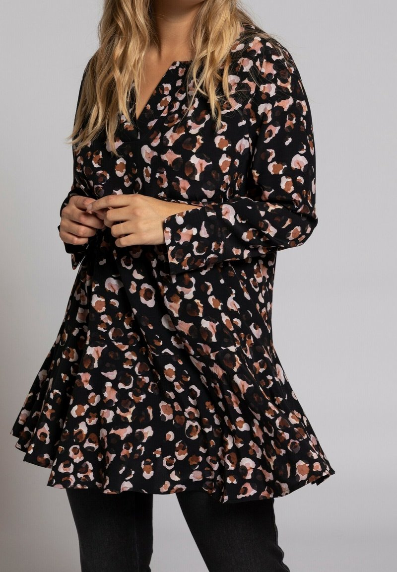 Kvinna som bär en lös svart tunika-klänning med ett brunt och rosa leopard-mönster, står med händerna vid midjan, mot en enkel ljus bakgrund.