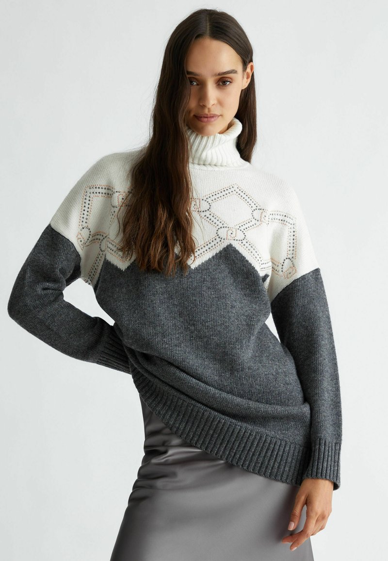 Maglione chunky con colletto alto; parte superiore bianca con dettagli a fantasia, corpo grigio scuro, polsini e orlo a costine, texture morbida.