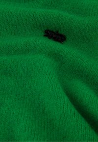 Groene brei stof met een zachte textuur heeft een klein zwart dierenembleem in de oppervlakte geweven.