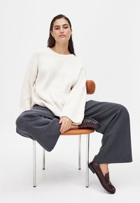 Weißer gerippter Pullover mit weiten Ärmeln, graue weite Hose und dunkelrote Loafer, sitzt auf einem braunen armleeren Stuhl vor einem weißen Hintergrund.