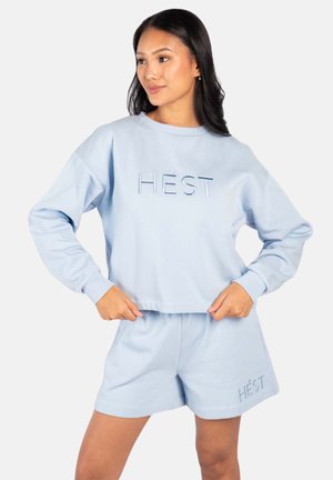 Kvinne iført lyseblå genser og matchende shorts med teksten "HÉST", står med hendene på hoftene og ser til siden.