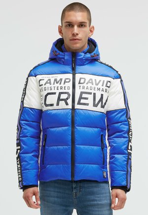 Blue puffer jacket with white and black accents, featuring large logos and text, zip closure, and contrasting side panels. Textured finish.