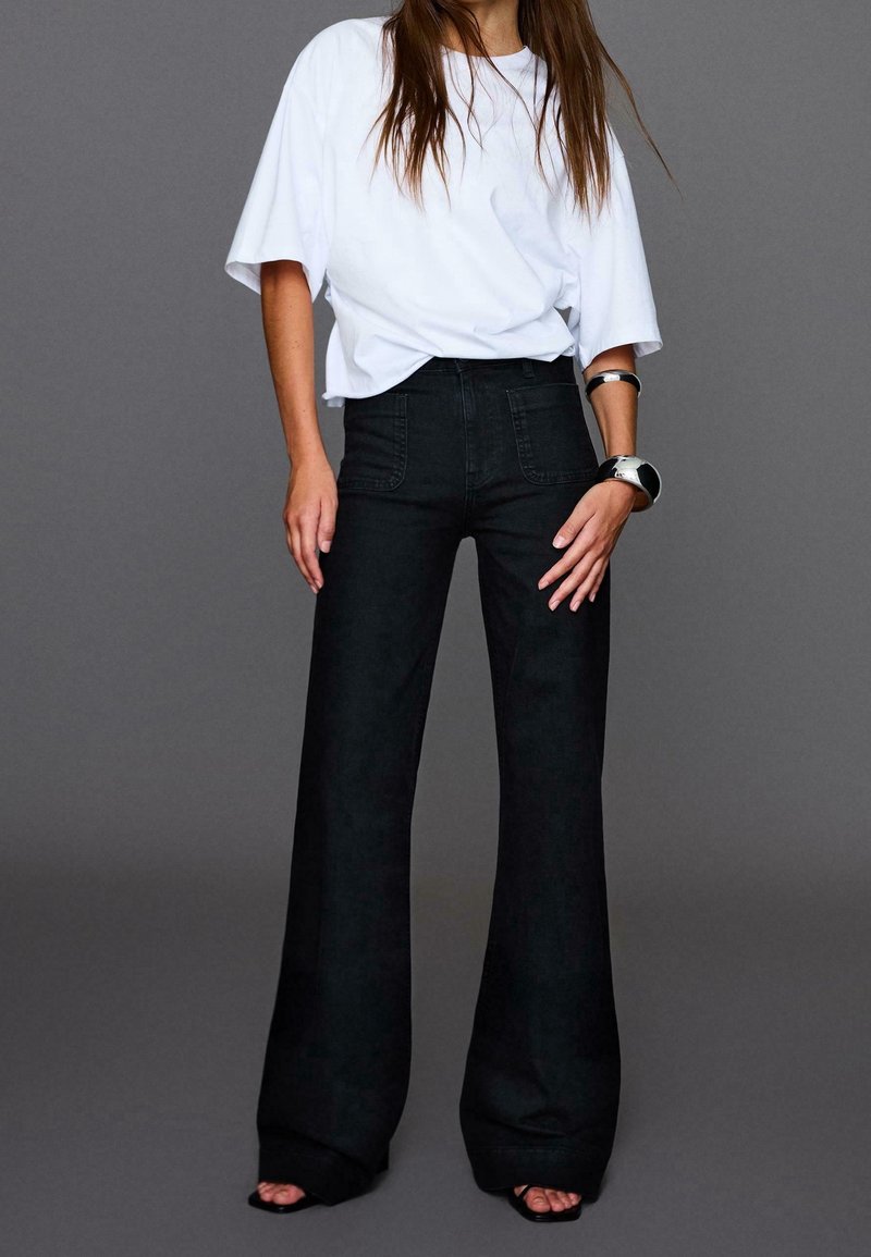 Femme portant un t-shirt blanc ample rentré dans un pantalon noir taille haute à jambes larges, avec des sandales noires et des bracelets argentés.