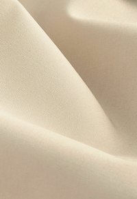Beige textiel met een gestructureerd oppervlak, met subtiele geweven patronen. Het materiaal valt soepel, waardoor er zachte plooien ontstaan.