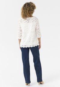 Top bianco in pizzo con maniche a tre quarti, orlo scallop, indossato sopra jeans blu scuro, mostrato da dietro. Texture liscia, motivo floreale.