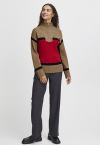 Der Strickpullover hat einen hohen Kragen, einen Reißverschluss, einen braunen Grundton, ein rotes Mittelteil und schwarze Streifen. Kombiniert mit grauen, maßgeschneiderten Hosen und schwarzen Schuhen.