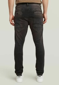 Άνδρας φορώντας ξεθωριασμένα μαύρα τζιν παντελόνια με διπλές πίσω τσέπες και λευκά sneakers, στέκεται μπροστά από μία απλή ανοιχτόχρωμη φόντο.