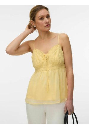 Femme portant un haut sans manches jaune avec des détails en dentelle, tenant un sac à main noir, sur fond blanc.