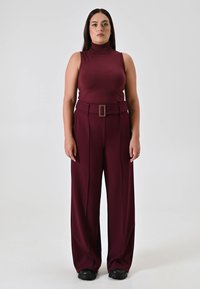 Haut sans manches en turtleneck bordeaux avec un design ajusté associé à un pantalon large taille haute avec une ceinture. Chaussures noires.