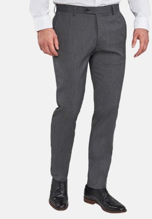 Homme portant un pantalon de costume gris, des chaussures habillées en cuir noir et une chemise blanche à manches longues, debout sur un fond blanc.