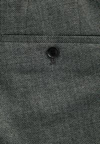 Tessuto grigio strutturato con un primo piano di un bottone nero. Il materiale presenta un motivo tessuto con leggere variazioni di ombreggiatura.