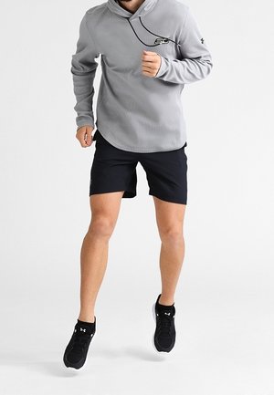 Uomo che corre indossando una maglietta a maniche lunghe grigia chiara, pantaloni corti neri e scarpe da corsa nere su uno sfondo bianco semplice.