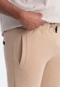 Pantaloni della tuta beige realizzati in tessuto morbido, dotati di una vita elastica con cordino, tasche laterali e una texture leggera e liscia.