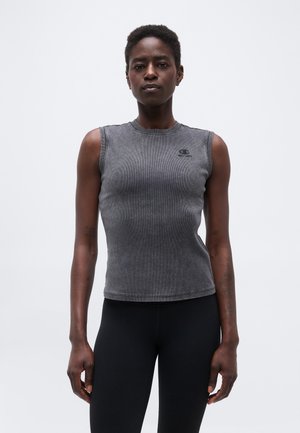 Žena v bezrukávovom priliehavom sivom tričku a čiernych nohaviciach stojaca čelom k divákovi na jednoduchom svetlom pozadí.