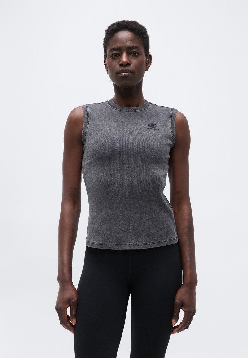 Γυναίκα που φοράει αμάνικο, εφαρμοστό γκρι τοπ και μαύρο παντελόνι, στέκεται μπροστά σε ένα απλό ανοιχτόχρωμο φόντο, κοιτάζοντας προς τα εμπρός.