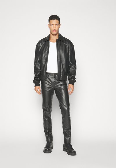 Svart läderbombarjacka med ribbade manschetter, parat med åtsittande svarta läderbyxor och svarta snörstövlar, mot en vit bakgrund.