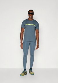 Miesten urheiluasu: lyhythihainen harmaa paita, jossa on neonkeltainen "DYNAFIT"-logo, vaaleanharmaat legginsit ja moniväriset juoksukengät.