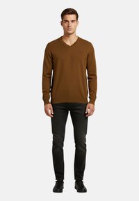 Maglione marrone con scollo a V in tessuto strutturato e maniche lunghe, abbinato a jeans neri e scarpe nere. Piccolo dettaglio con logo sul maglione.