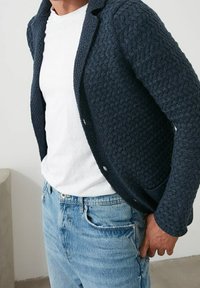 Cardigan de punto en color marino con un patrón texturizado, que presenta un cuello con muesca y cierre de botones, llevado sobre una camiseta blanca y jeans de mezclilla claros.