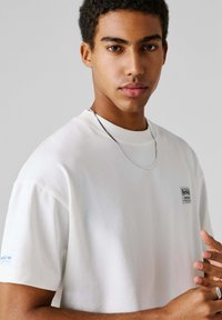 Λευκό βαμβακερό t-shirt με κοντά μανίκια, που διαθέτει ένα μικρό μπαλώμα με το λογότυπο στο στήθος, απλός σχεδιασμός, στρογγυλή λαιμόκοψη και λεία υφή.
