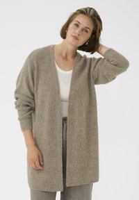 Femme avec les cheveux bruns courts portant de grandes boucles d'oreilles en or, un long cardigan beige, un top blanc et un pantalon marron clair, posant avec une main sur la tête.