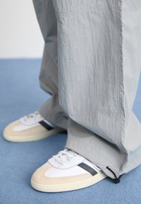 Bílé a béžové tenisky s černými pruhy obuté na volných světle šedých kalhotách na modré podlaze.