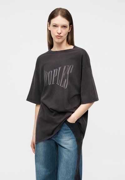 Νεαρή γυναίκα με ίσια καστανά μαλλιά που φοράει μια φαρδιά μαύρη μπλούζα με το κείμενο "PEOPLES" και δίχρωμο τζιν μπλε, με το χέρι στην τσέπη.