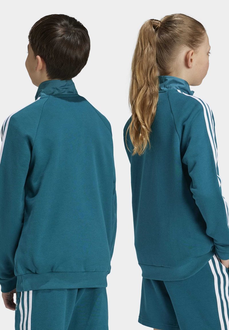 Chaqueta de manga larga de color verde azulado con acentos de rayas blancas, puños y dobladillo acanalados. Tejido suave, diseñado para la comodidad. Vista desde la parte de atrás, usada por dos niños.
