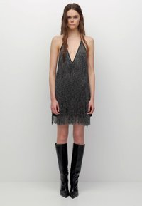 Robe à franges noire avec un décolleté en V profond, réalisée en fil métallique. Présente une coupe ample et s'arrête juste au-dessus du genou, portée avec des bottes hautes jusqu'au genou.