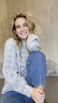 Model draagt een grijs geruit hemd en blauwe spijkerbroek, zit met opgetrokken knieën en lacht tegen een gestructureerde grijze muur.