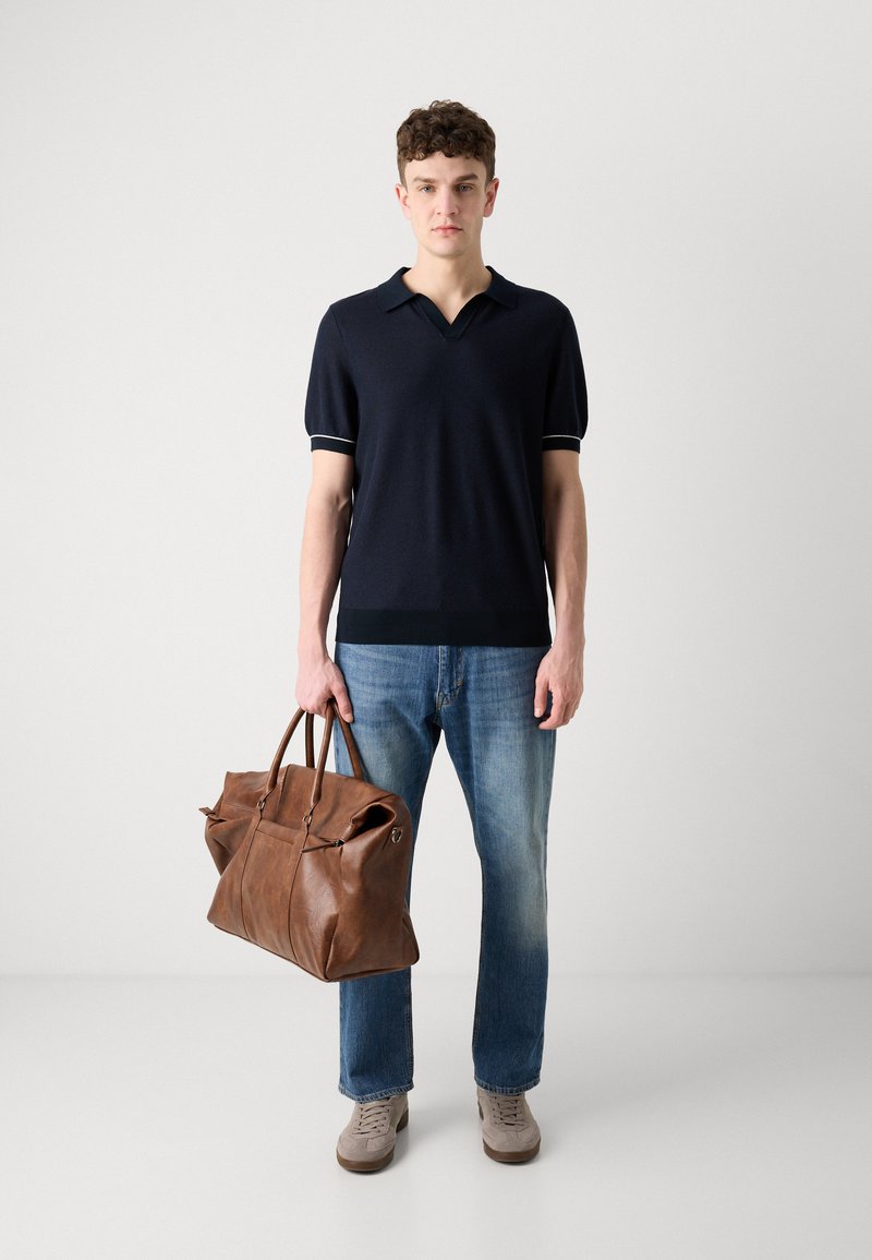 Polo bleu marine à manches courtes avec un col en V et des accents clairs, associé à un jean bleu et un grand sac de voyage en cuir marron. Baskets grises.