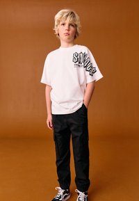 Weißes bedrucktes T-Shirt mit dem Schriftzug "SAVAGE NEW YORK CITY", kombiniert mit schwarzen Cargo-Hosen und schwarz-weißen Turnschuhen, vor braunem Hintergrund.
