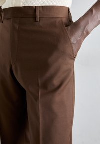 Bruna skräddarsydda byxor med en slät yta, som har sidofickor och en rak passform, vilket framhäver en modern design.