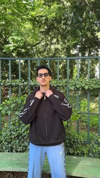 Homme portant des lunettes de soleil, un sweat à capuche noir et un pantalon bleu clair, debout devant une clôture métallique verte avec du lierre et des arbres derrière.
