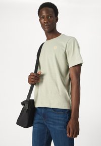 Maglietta di cotone verde chiaro con scollatura tonda e maniche corte, con una piccola grafica di un fiore. Abbinata a jeans in denim blu e a una borsa nera.