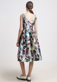 Vestido midi floral con un patrón de pensamientos coloridos, diseño sin mangas, falda más amplia y espalda descubierta, hecho de tela brillante. Combinado con zapatos blancos.