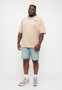 Beżowa bawełniana koszulka z krótkim rękawem i logo "Tropica", w połączeniu z jasnoniebieskimi szortami dżinsowymi. Szare sneakersy z białymi akcentami.
