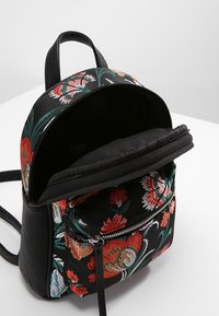 Czarny plecak materiałowy z kolorową haftowaną aplikacją kwiatową, zamykaną kieszenią z przodu oraz teksturowanym wykończeniem. Otwarta góra odsłania czarne wnętrze.