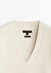 Pull à col en V de couleur blanc cassé, présentant une texture côtelée et des motifs en maille torsadée. L'étiquette indique "Fabriqué en Turquie", précisant le pays de fabrication.