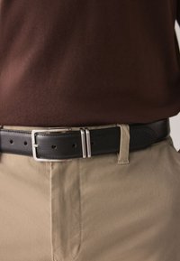 Black leather belt with a silver metal buckle, featuring multiple holes and two sleek silver accents, worn over beige pants.