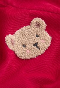 Il tessuto rosso presenta un ricamo testurizzato di un volto di orso in beige con dettagli neri sugli occhi e sul naso, e accenti rosa sulle guance.